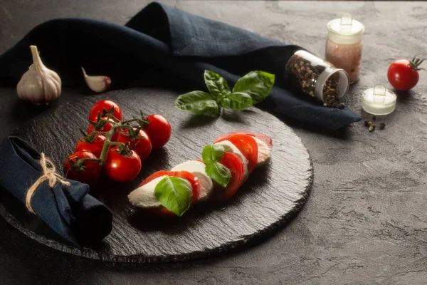 Salada Caprese em prato de pedra com mussarela, folhas de manjericão fresco, tomates, sal marinho, pimenta seca e alho fresco no fundo escuro . — Fotografia de Stock