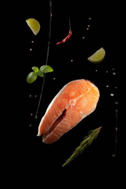 Fesleğen yapraklı çiğ somon bifteği, taze biberiye, kırmızı biber, limon dilimleri, baharatlar, deniz tuzu ve siyah bir zeminde biraz yağ..