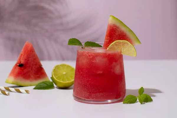 Cocktail all'anguria in un bicchiere con foglie di menta, lime e cubetti di ghiaccio su fondo viola con ombre di foglie di palma.Cocktail freddo rinfrescante o frullato. — Foto Stock