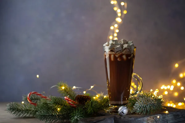 Gorąca czekolada lub kakao z piankami i cynamonem w proszku z gałązkami jodły na drewnianej desce vintage z lampkami świątecznymi, cukierkami, dzwoneczkami.. — Zdjęcie stockowe