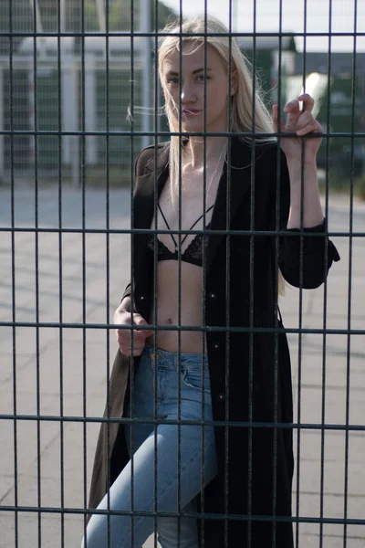 金属フェンスの後ろに黒いコートでブロンドの女の子 — ストック写真