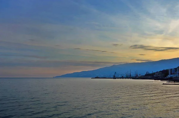 Denize, gün batımı gökyüzüne, dağa, sahildeki şehir ve crans siluetleri ile liman manzarası