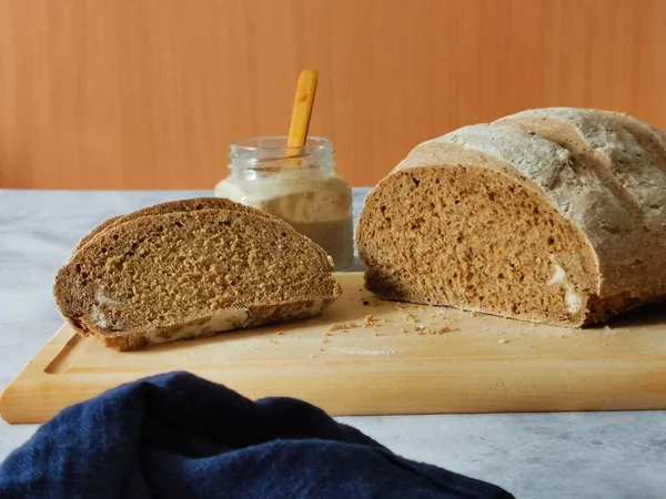 Cortar el pan de centeno en una tabla de madera y un frasco de masa madre sobre el fondo de una pared de madera —  Fotos de Stock