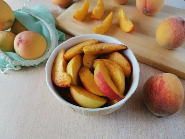 juicy sliced peaches in a plate, next to whole peaches in a reusable bag and on a wooden plank