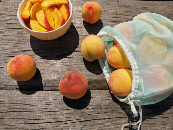 juicy sliced peaches in a plate, whole peaches and a reusable bag on old wooden table. bright sun, hard shadows