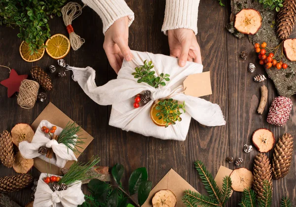 Christmas Zero waste, eco friendly packaging. Woman is wrapping gifts in linen fabric on dark rustic wooden background. Ecological Christmas holiday concept, eco decor.