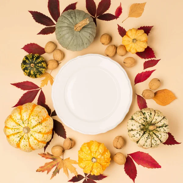 Autumn fall holiday Thanksgiving table place setting. Pumpkins, porcelain white plate, dry maple leaves, spice and apple on beige pastel background. Flat lay, top view, copy space.