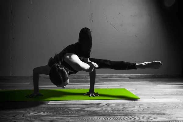 Foto en blanco y negro de Joven hermosa mujer morena en ropa negra practica yoga Parivritta Kaundiniasana en estudio oscuro estera verde Meditación deportiva monocromo — Foto de Stock