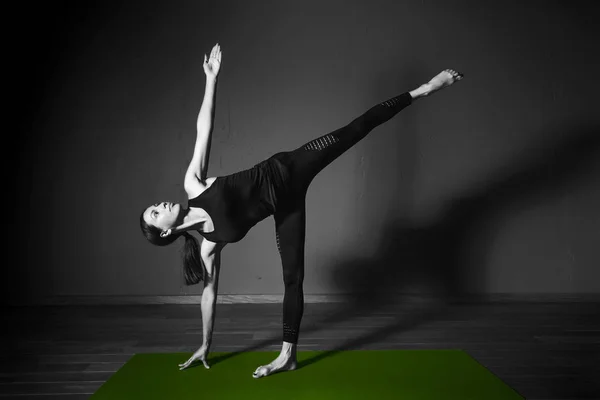 Black white photo of Young beautiful woman brunette in black clothes practice yoga ardha chandrasana in dark studio green mat Sport meditation monochrome — Stock Photo, Image