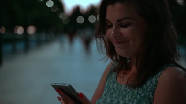 夕暮れ時に公園の青いドレスにピンクの爪を持つ美しい女性は笑顔で スマートフォンでチャットやテキストを笑い 友人や家族と連絡を取り合っていますスローモーション — ストック動画