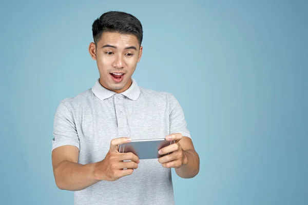 Азиатский молодой красивый привлекательный мужчина играет в игру со смартфоном на синем фоне — стоковое фото