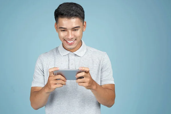 Asian młody przystojny atrakcyjny człowiek gra z inteligentny telefon na niebieskim tle — Zdjęcie stockowe