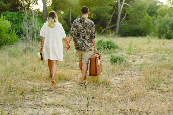 Heterosexual couple walking through countryside with a suitcase to travel and a hat