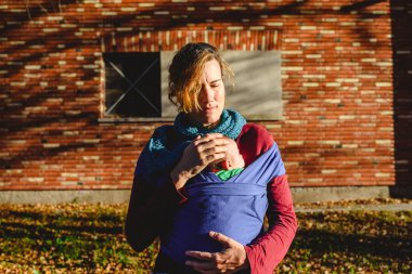 sevgiyle kırmızı tuğla bina önünde bir gün batımı yürüyüş sırasında bir eşarp yeni doğan bebeğini taşıyan anne.