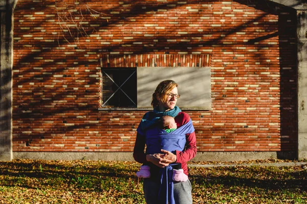 sevgiyle kırmızı tuğla bina önünde bir gün batımı yürüyüş sırasında bir eşarp yeni doğan bebeğini taşıyan anne.