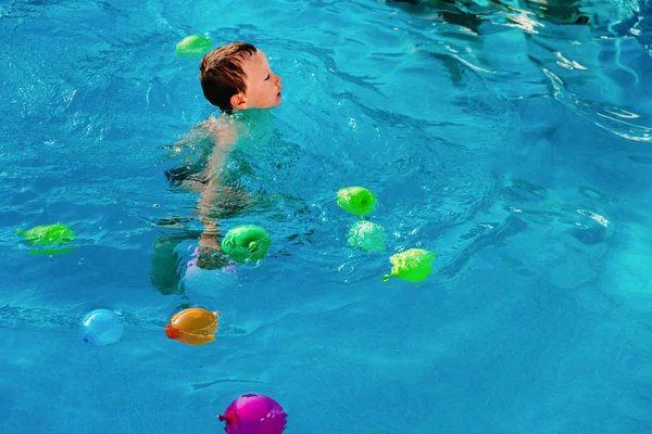 Young blond boy playing in a pool with water balloons, colorful, — Stock Photo, Image