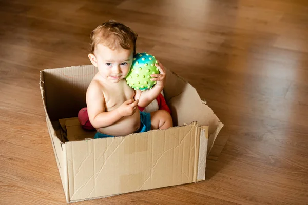 Niña jugando con una pelota dentro de una caja de cartón y haciendo — Foto de Stock