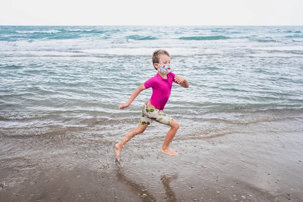 A boy runs along the shore of the beach wearing a face mask during the new normality.