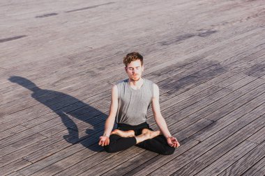 Genç kızıl saçlı adam nilüfer yogası yapıyor, Padmasana, açık havada, ahşap zeminde.