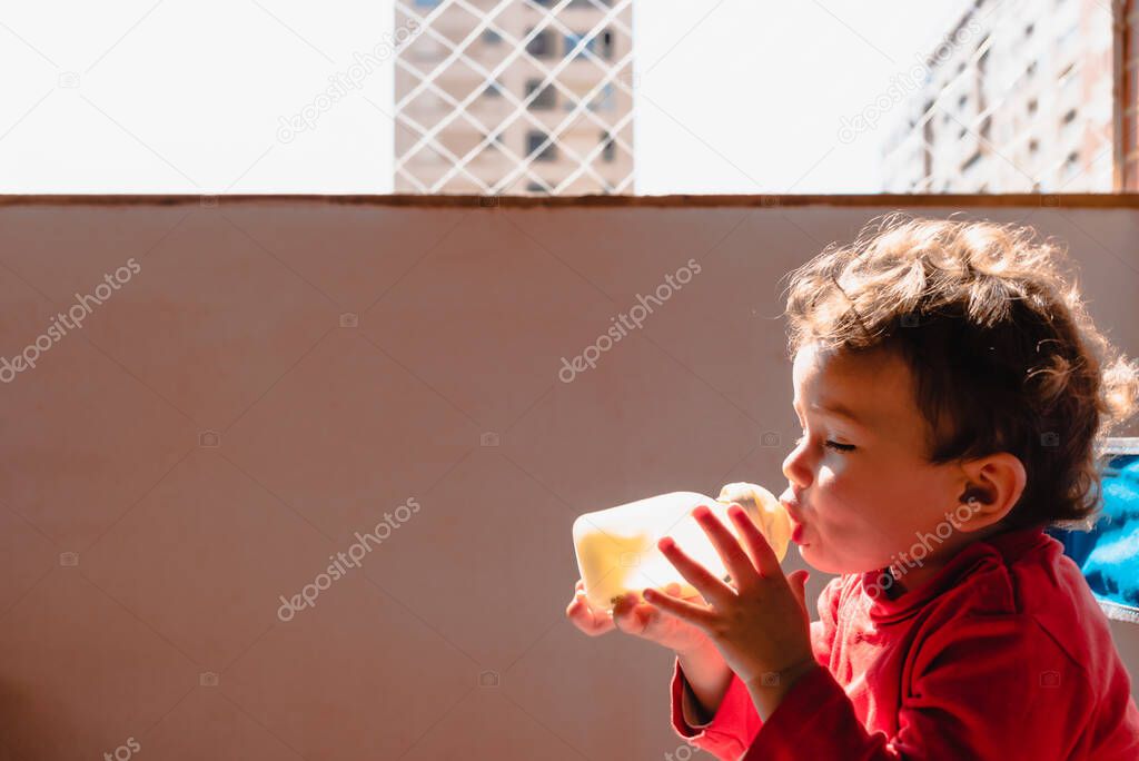 Baby sitting on the balcony of her house in the sun drinks milk from a bottle by herself.