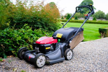 Sağlam bir çim biçme makinesi, kırmızı ve gri tasarımını sergileyen çakıllı bir yolda dinleniyor. Açık hava bakımını vurgulayan yeşil çitler ve sakin manzaralarla çerçevelenmiştir..