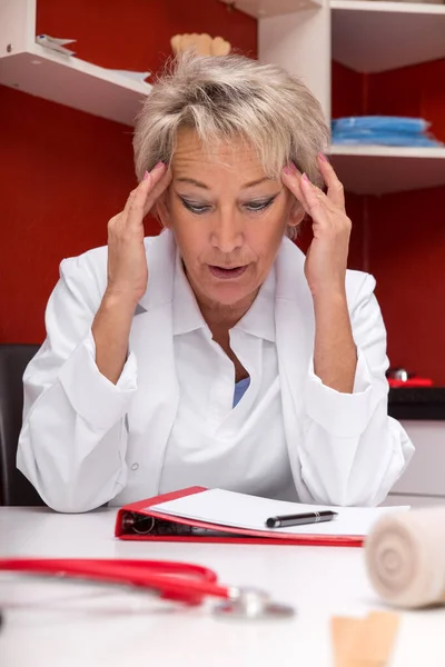 头痛的老年女医生 也许累坏了 桌子上的文件 红色办公室 — 图库照片