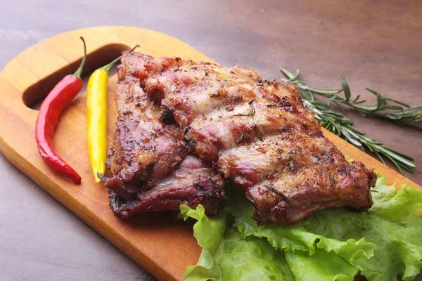 Grilled barbecued ribs with lettuce leaves, hot chili pepper and sauce on wooden cutting board. — Stock Photo, Image