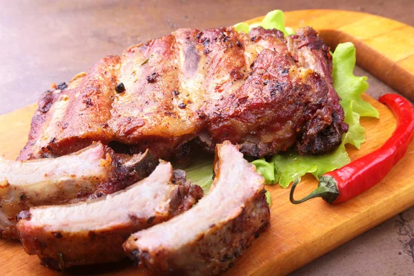 Grilled barbecued ribs with lettuce leaves, hot chili pepper and sauce on wooden cutting board. — Stock Photo, Image