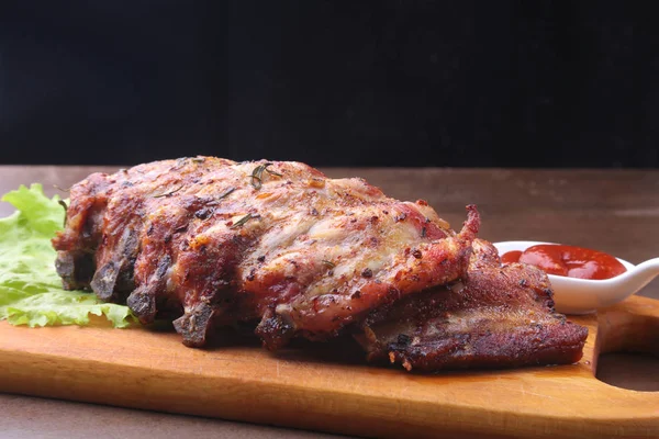 Grilled barbecued ribs with lettuce leaves, hot chili pepper and sauce on wooden cutting board. — Stock Photo, Image