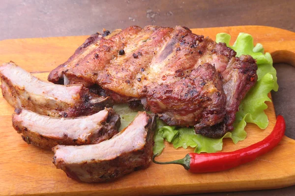 Grilled barbecued ribs with lettuce leaves, hot chili pepper and sauce on wooden cutting board. — Stock Photo, Image