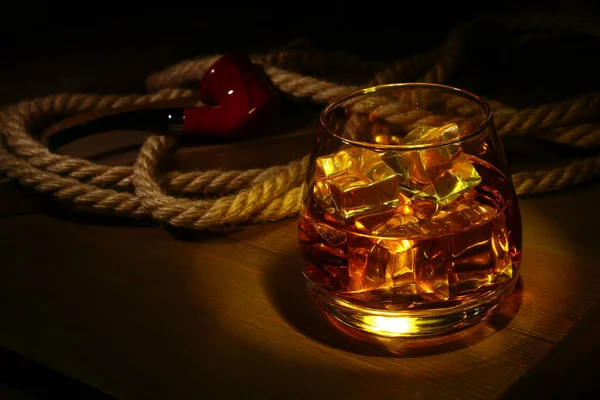 Glass of whiskey, cognac or brandy with ice and smoking pipe on wood table. Top view with copy space — Stock Photo, Image