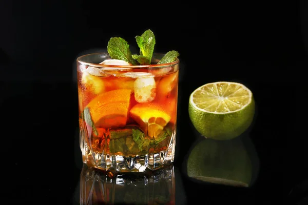 Bicchiere di Rum scuro Cocktail con lime, arancia, cubetti di ghiaccio e foglie di menta su fondo nero a specchio . — Foto Stock