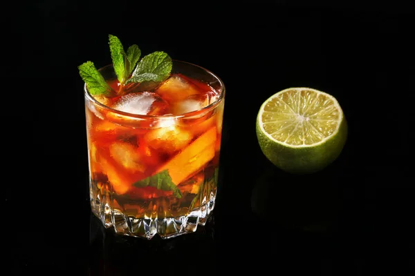 Vaso de ron oscuro Cóctel con lima, naranja, cubitos de hielo y hojas de menta sobre fondo de espejo negro . — Foto de Stock