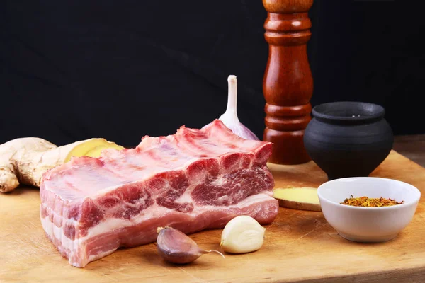 Raw ribs with a garlic, spices, ginger, salt and grinder on cutting board. Black background. — Stock Photo, Image