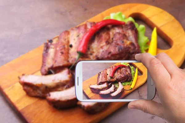 Manos del hombre con teléfono inteligente tomando fotos asadas costillas a la parrilla con hojas de lechuga, chile picante y salsa en la tabla de cortar de madera . —  Fotos de Stock