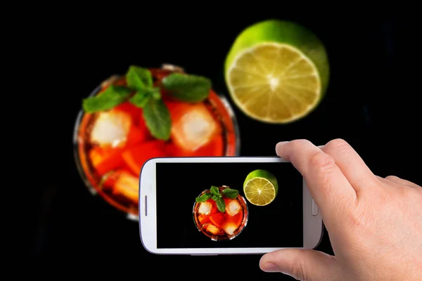 Ręce człowieka ze smartfonem biorąc zdjęcie szklanka ciemnego rumu koktajl z limonką, pomarańczą, kostki lodu i liści mięty na czarnym tle lustro. — Zdjęcie stockowe
