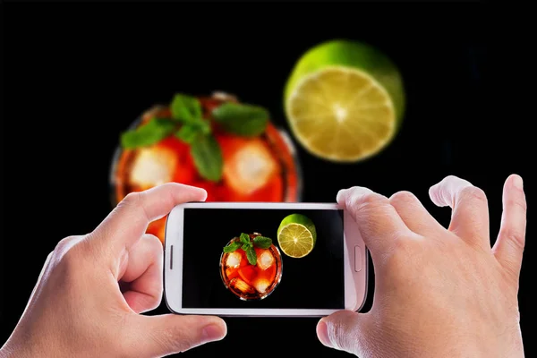 Ręce człowieka ze smartfonem biorąc zdjęcie szklanka ciemnego rumu koktajl z limonką, pomarańczą, kostki lodu i liści mięty na czarnym tle lustro. — Zdjęcie stockowe