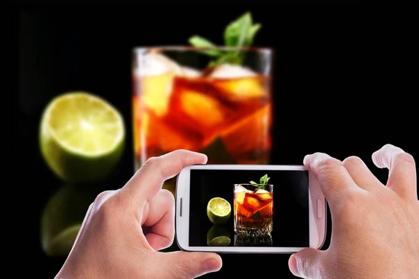 Manos del hombre con teléfono inteligente tomando la foto Vaso de ron oscuro Cóctel con lima, naranja, cubitos de hielo y hojas de menta sobre fondo de espejo negro . —  Fotos de Stock