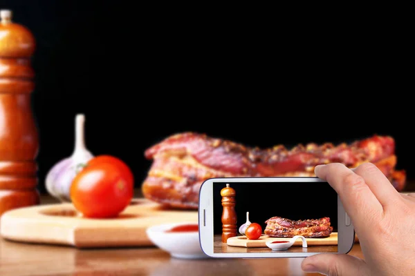Ręce człowieka ze smartfonem robienia zdjęć grillowane żebra wieprzowe Grill Striploin stek z sosem Chimichurri i pomidorami na płycie do krojenia na ciemnym tle. — Zdjęcie stockowe
