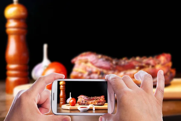 Ręce człowieka ze smartfonem robienia zdjęć grillowane żebra wieprzowe Grill Striploin stek z sosem Chimichurri i pomidorami na płycie do krojenia na ciemnym tle. — Zdjęcie stockowe