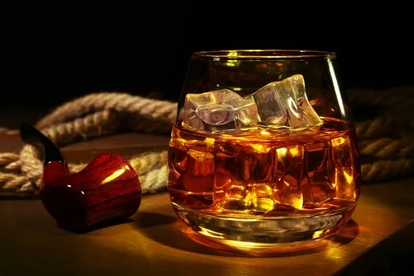 Glass with whiskey, ice cubes and smoking pipe on wooden background. — Stock Photo, Image