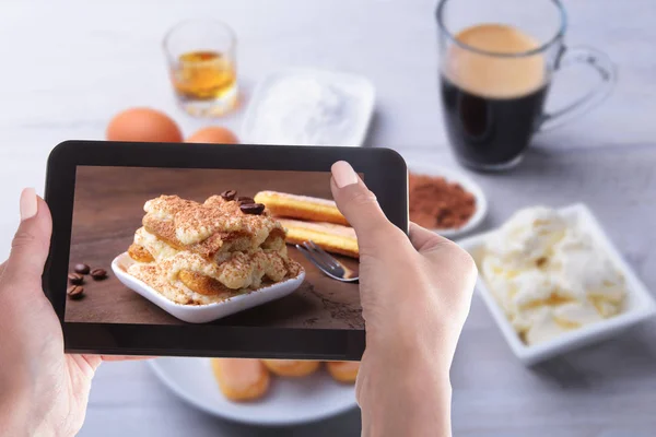 Manos femeninas con un teléfono inteligente, en la pantalla de la cual hay una foto del pastel de tiramisú en el fondo de los ingredientes para el pastel . —  Fotos de Stock