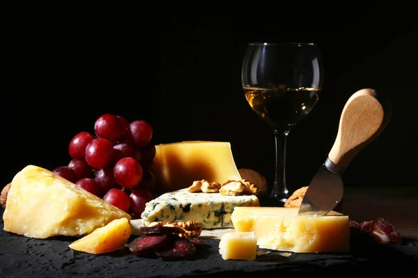 Assorted cheeses, nuts, grapes, fruits, smoked meat and a glass of wine on a serving table. Dark and Moody style. Free space for text. — Stock Photo, Image