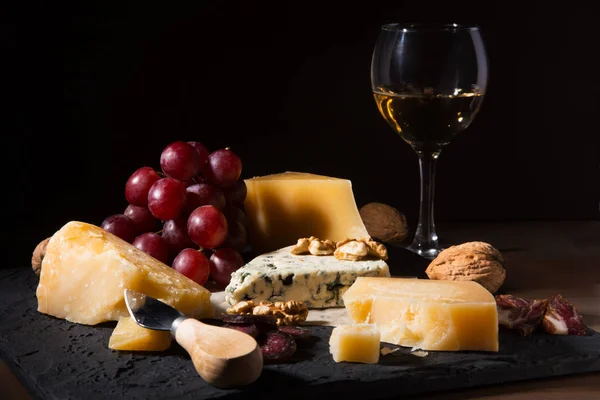 Quesos variados, frutos secos, uvas, frutas, carne ahumada y una copa de vino en una mesa de servir. Estilo oscuro y Moody. Espacio libre para texto . — Foto de Stock