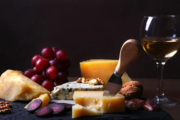 Quesos variados, frutos secos, uvas, frutas, carne ahumada y una copa de vino en una mesa de servir. Estilo oscuro y Moody. Espacio libre para texto . — Foto de Stock