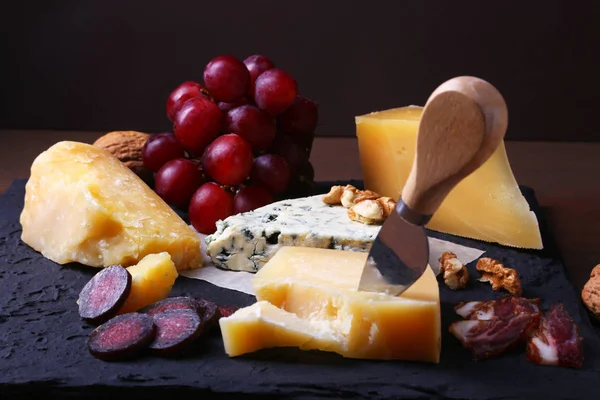 Diverse ostar, nötter, vindruvor, frukter, rökt kött och ett glas vin på ett serveringsbord. Mörk och lynnig stil. Ledigt utrymme för text. — Stockfoto