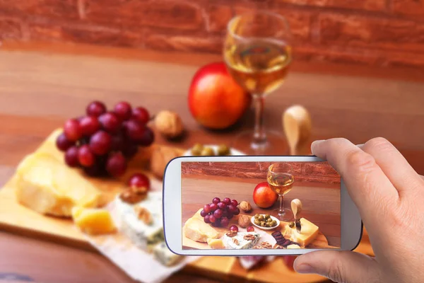 Manos del hombre con el teléfono inteligente tomar fotos Surtido de quesos, frutos secos, uvas, frutas, carne ahumada y una copa de vino en una mesa de servir. Estilo oscuro y Moody . —  Fotos de Stock