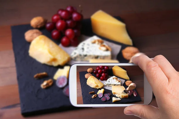 Manos del hombre con el teléfono inteligente tomar fotos Surtido de quesos, frutos secos, uvas, frutas, carne ahumada y una copa de vino en una mesa de servir. Estilo oscuro y Moody . —  Fotos de Stock