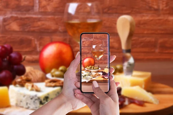 Mani di donna con smartphone scattare foto Formaggi assortiti, noci, uva, frutta, carne affumicata e un bicchiere di vino su un tavolo da portata. Scuro e stile Moody . — Foto Stock