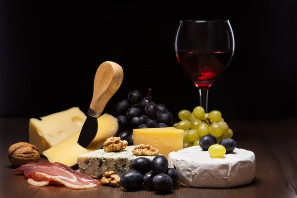 Queijos variados, nozes, uvas, frutas, carne defumada e um copo de vinho em uma mesa de serviço. Estilo escuro e mal-humorado. Espaço livre para texto . — Fotografia de Stock